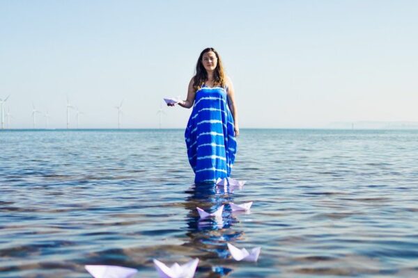 Lisette Auton standing in the sea with a lie of folded paper boats in front of her. She is wearing a blue and white dress.